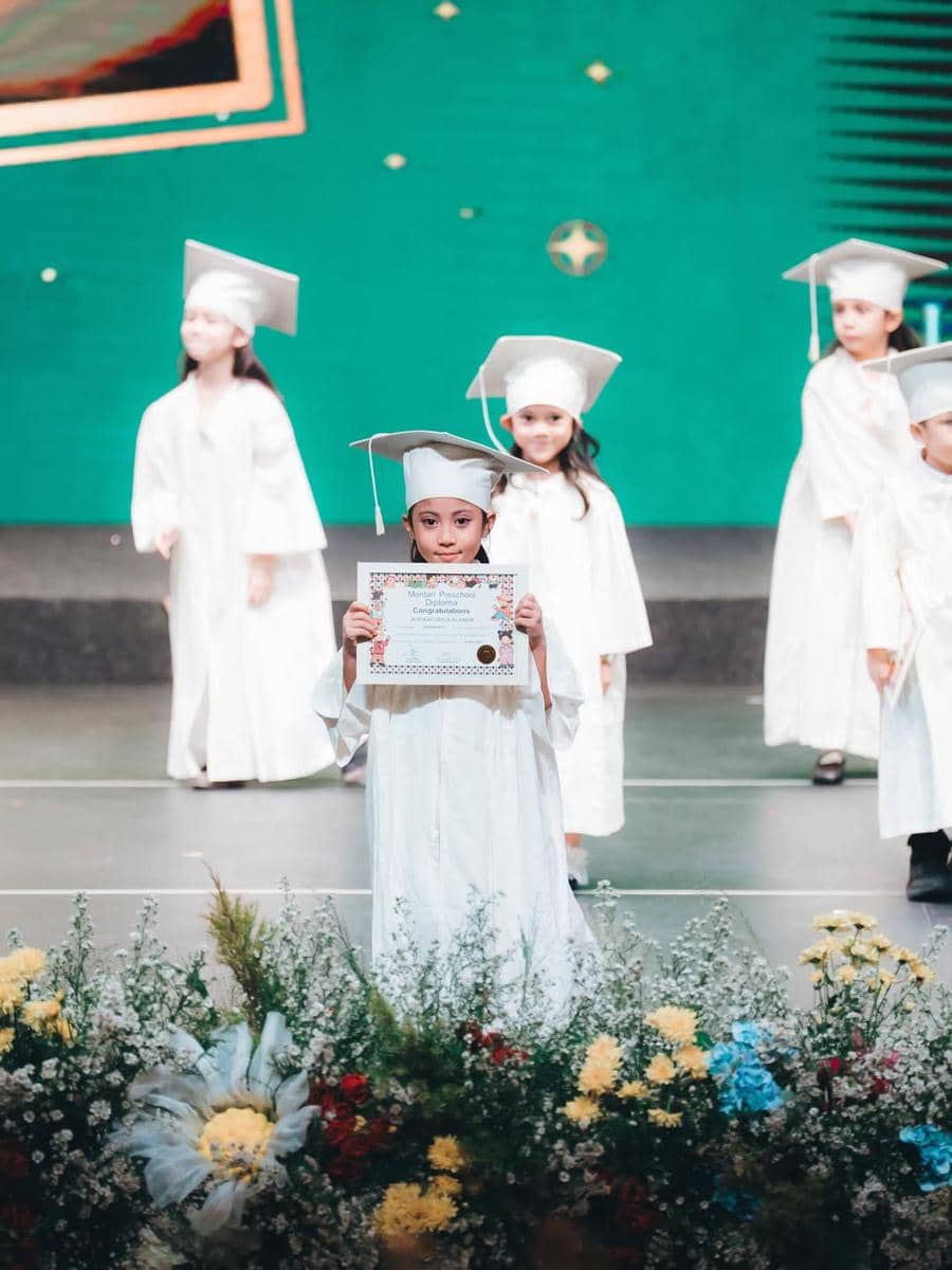 Seorang anak mengenakan toga putih memegang ijazah di atas panggung wisuda bersama teman-temannya dengan latar dekorasi hijau.