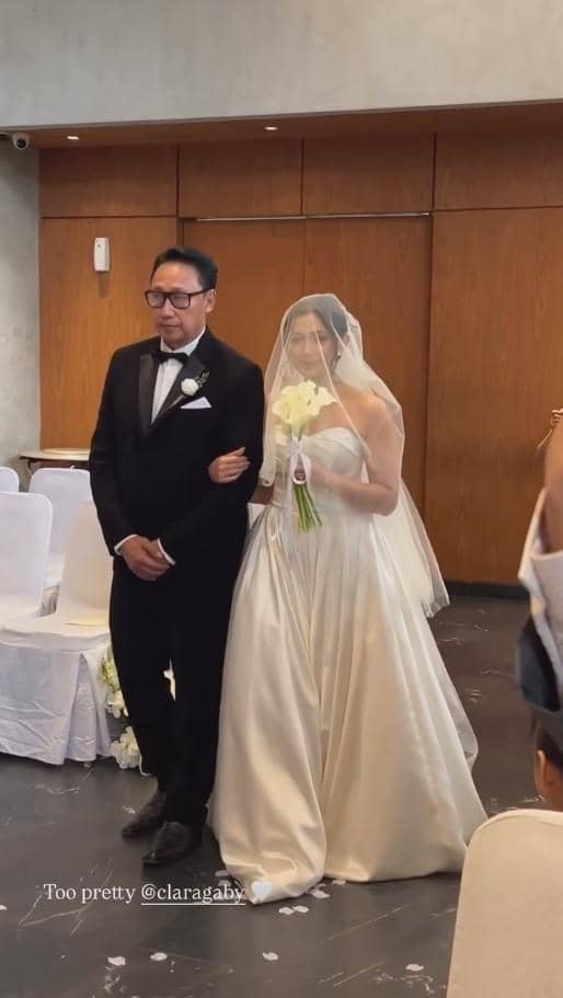 Seorang pengantin wanita mengenakan gaun putih dan kerudung berjalan di lorong pernikahan didampingi pria berjas hitam di sebuah ruangan.