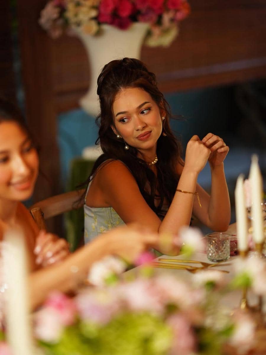 Seorang perempuan mengenakan gaun pastel duduk di meja makan berhias bunga dan lilin, tersenyum hangat kepada temannya.