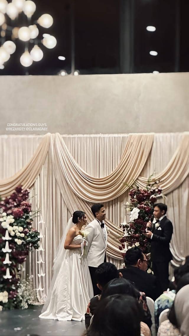 Pasangan pengantin berdiri di pelaminan dengan dekorasi bunga dan tirai krem, mendengarkan ucapan dari seorang pria berpakaian jas hitam.