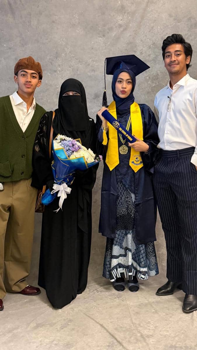 Seorang wisudawati mengenakan toga biru berpose bersama tiga anggota keluarga, salah satunya membawa buket bunga.