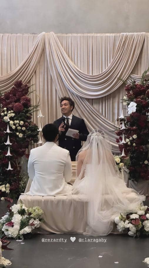 Pasangan pengantin duduk di depan penghulu saat upacara pernikahan dengan dekorasi bunga dan tirai elegan berwarna krem.