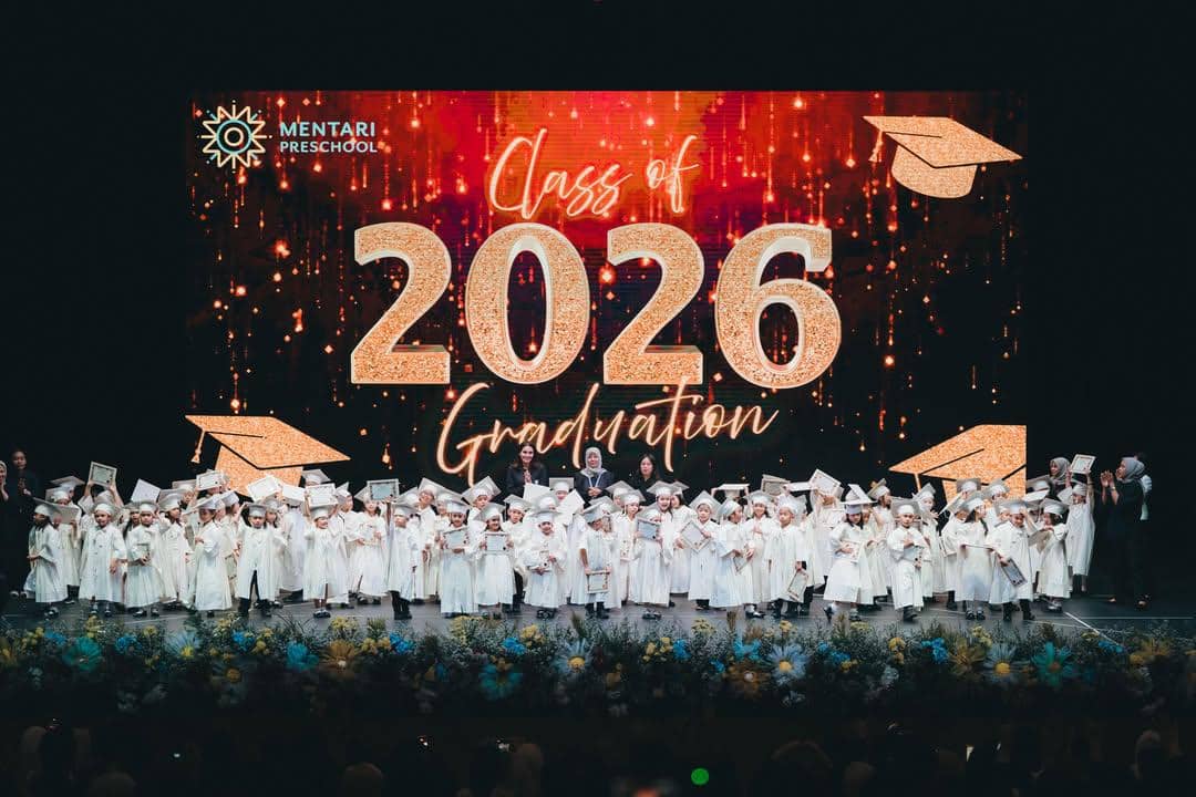 Anak-anak mengenakan toga putih berdiri di panggung saat wisuda TK Mentari Preschool dengan latar tulisan Class of 2026 Graduation.