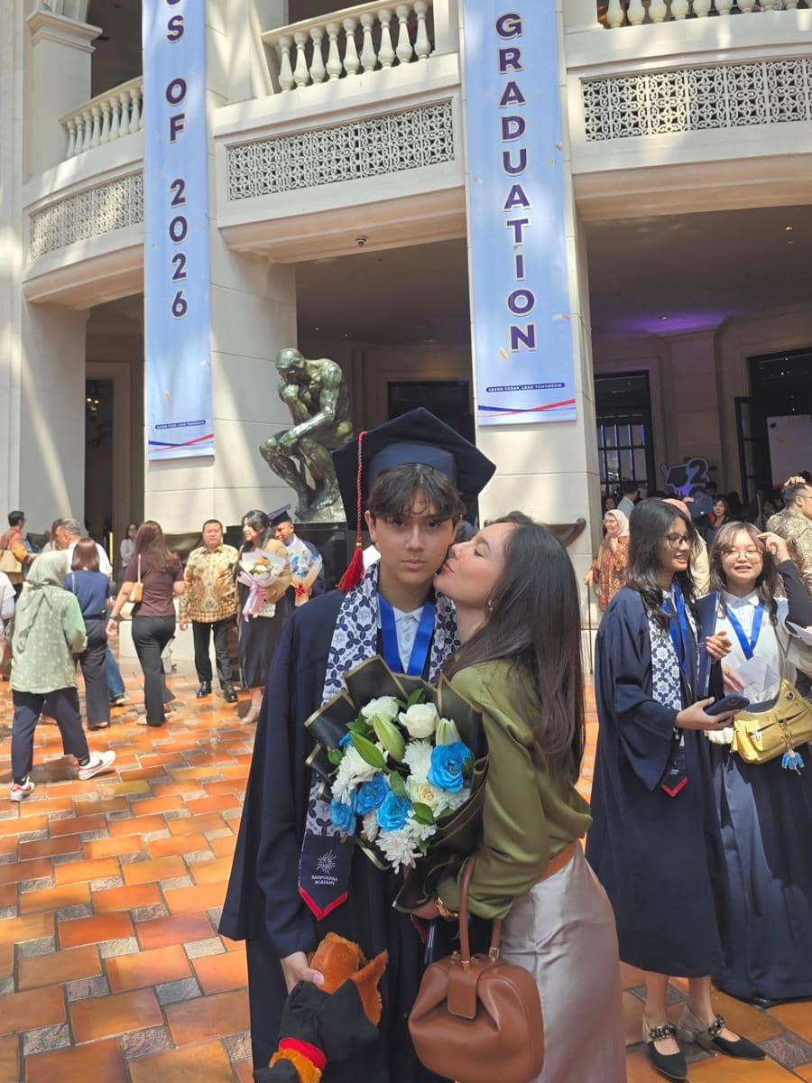 Seorang siswa mengenakan toga wisuda memegang buket bunga biru dan putih, dicium oleh seorang wanita di acara kelulusan tahun 2026.