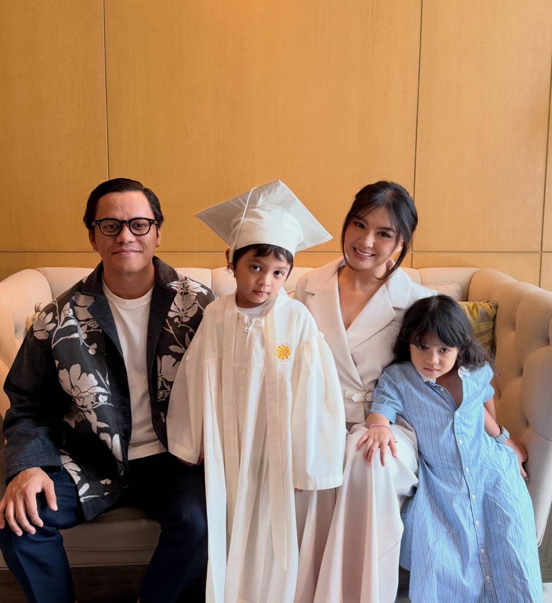 Seorang anak mengenakan toga wisuda putih berfoto bersama orang tua dan saudara di atas sofa dengan latar dinding kayu.