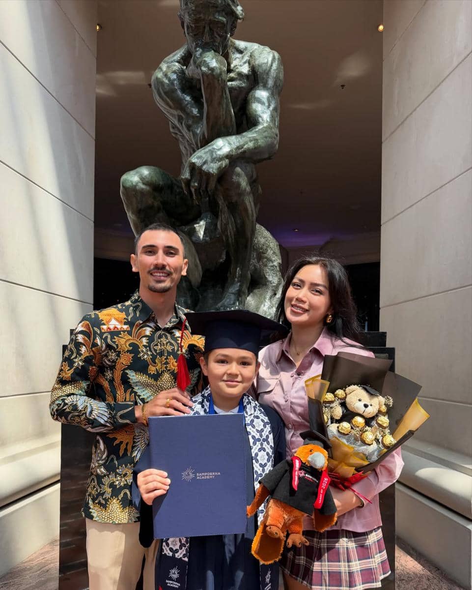 Seorang anak mengenakan toga wisuda berpose bersama dua orang dewasa sambil memegang ijazah dan buket hadiah di depan patung besar.