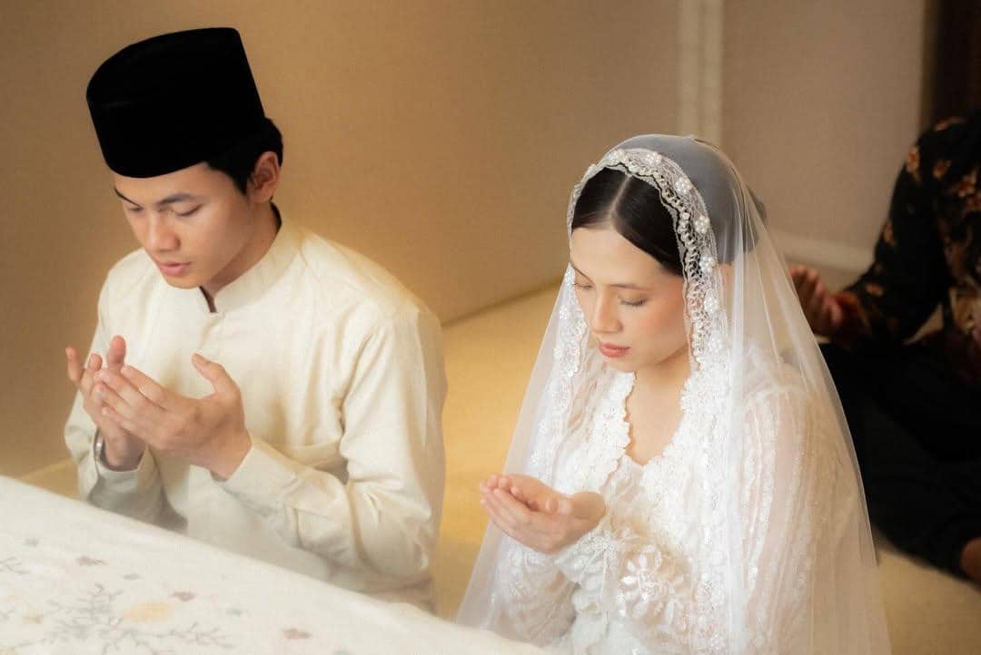 Sepasang pengantin mengenakan busana adat putih sedang berdoa bersama dalam suasana khidmat di hari pernikahan mereka.