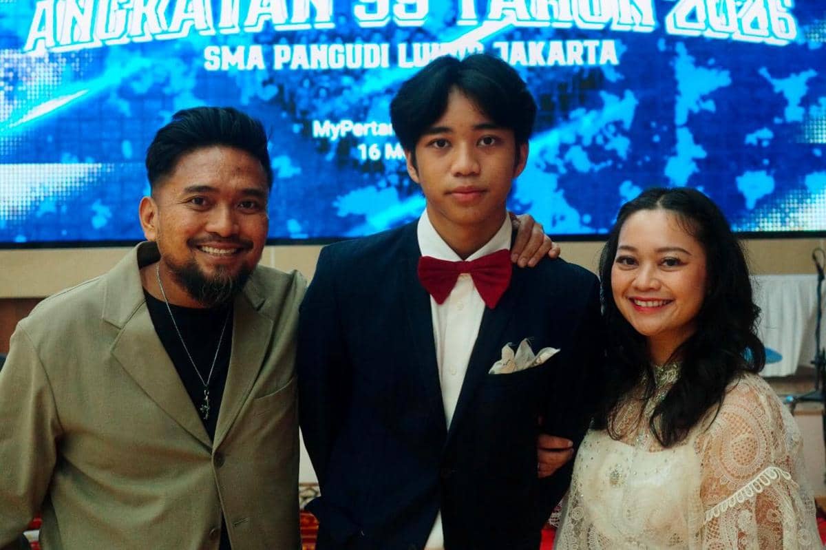 Seorang siswa SMA mengenakan setelan jas dengan dasi kupu-kupu merah berfoto bersama dua orang dewasa di acara wisuda sekolah tahun 2026.