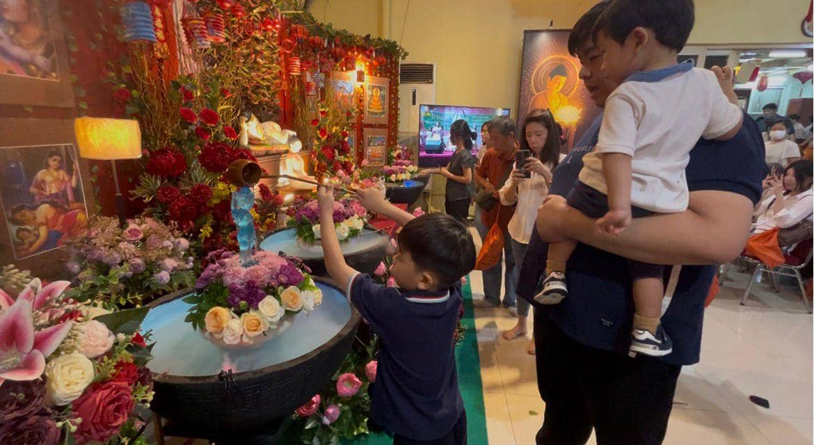 Seorang anak melakukan prosesi memandikan patung Buddha kecil saat perayaan Waisak, didampingi keluarga di sebuah vihara yang dihiasi bunga.
