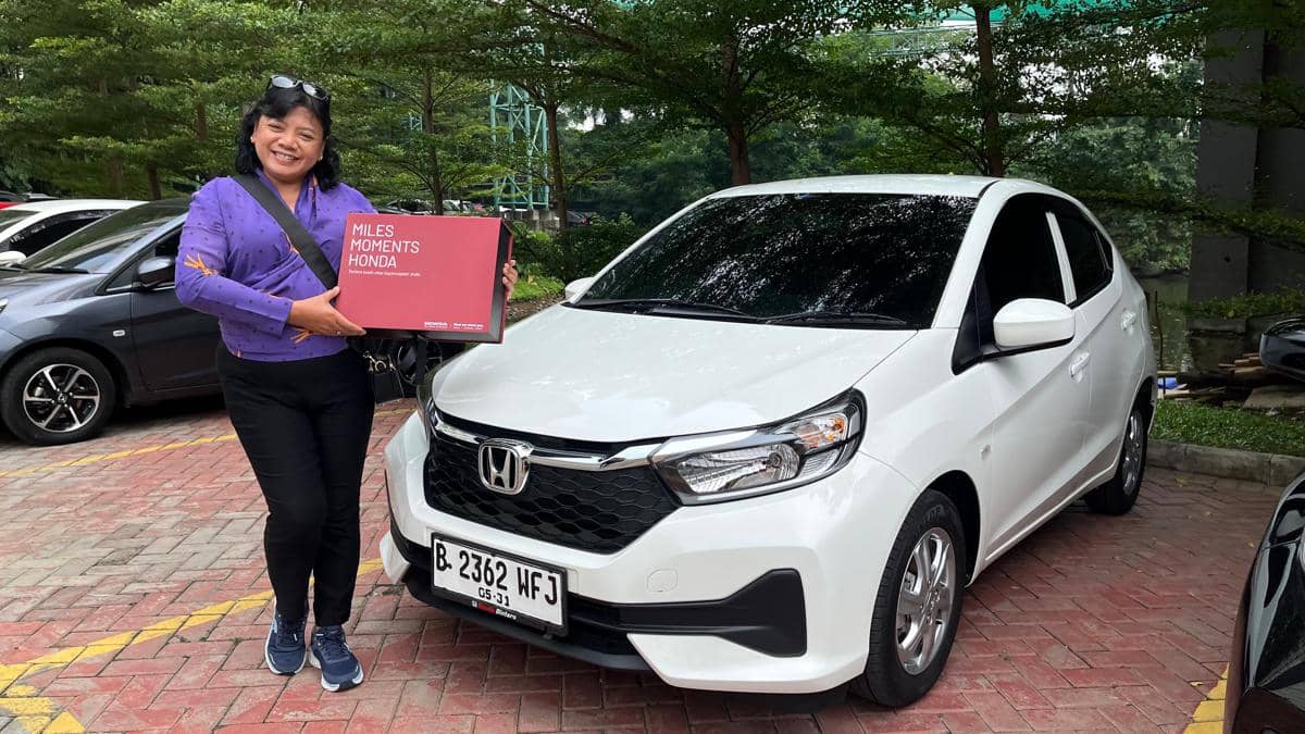 Seorang wanita berpose sambil memegang gift box bertuliskan Miles Moments Honda di samping mobil Honda Brio putih di area parkir.