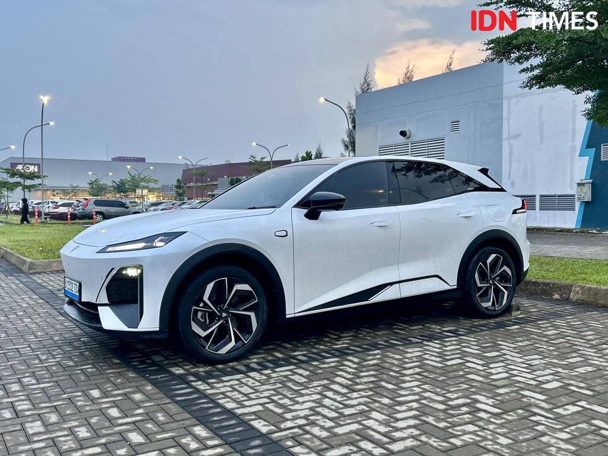 Mobil SUV listrik berwarna putih diparkir di area luar dengan latar gedung dan langit senja di belakangnya.