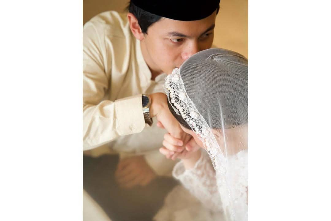 Seorang pria mengenakan pakaian tradisional berwarna krem mencium tangan wanita berhijab dengan gaun renda putih dalam suasana pernikahan.