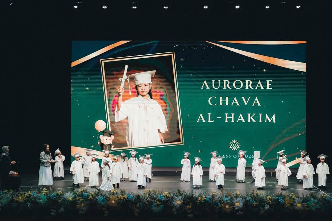 Anak-anak mengenakan toga putih mengikuti wisuda taman kanak-kanak di atas panggung dengan layar besar menampilkan nama Aurorae Chava Al-Hakim.