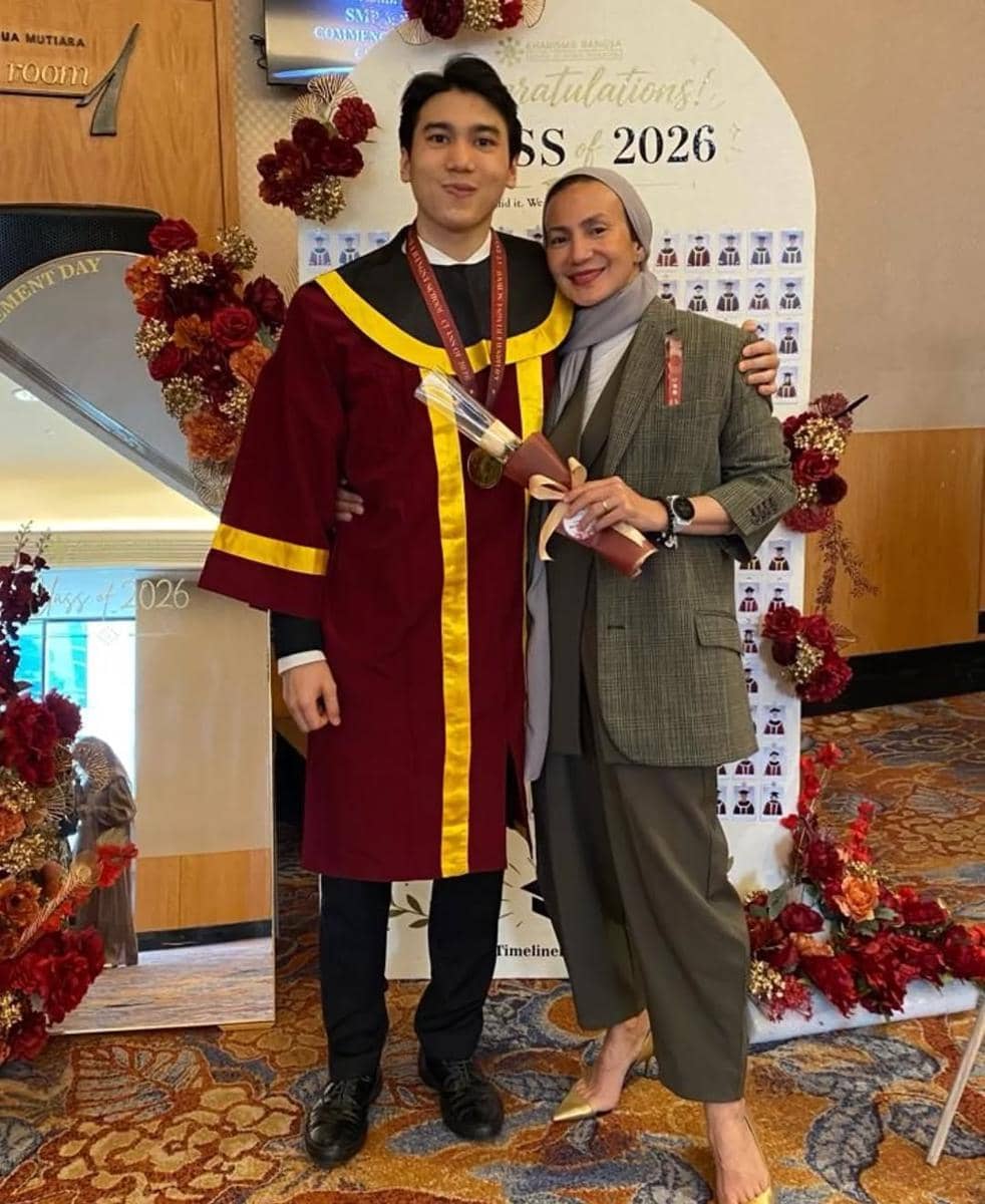 Seorang lulusan mengenakan jubah wisuda merah marun berfoto bersama seorang wanita berhijab di acara kelulusan tahun 2026.