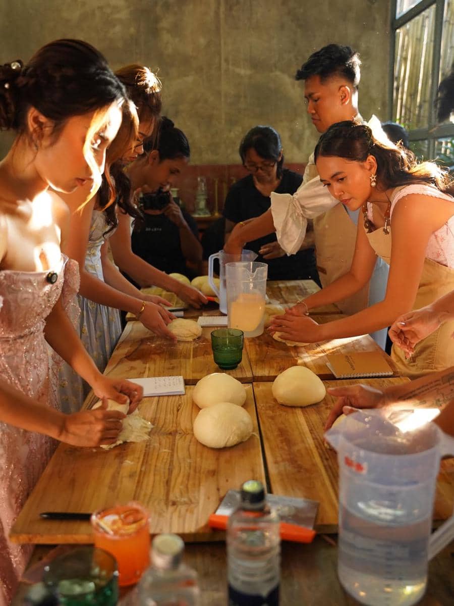 Beberapa wanita mengenakan gaun pesta dan celemek sedang menguleni adonan roti di meja kayu panjang dengan cahaya sore masuk dari jendela.