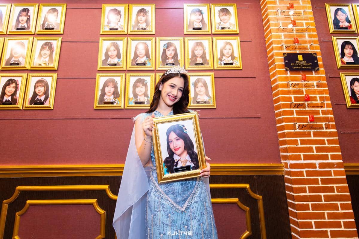 Seorang anggota JKT48 mengenakan gaun biru dan mahkota sambil memegang bingkai foto dirinya di depan dinding berisi potret anggota lain.