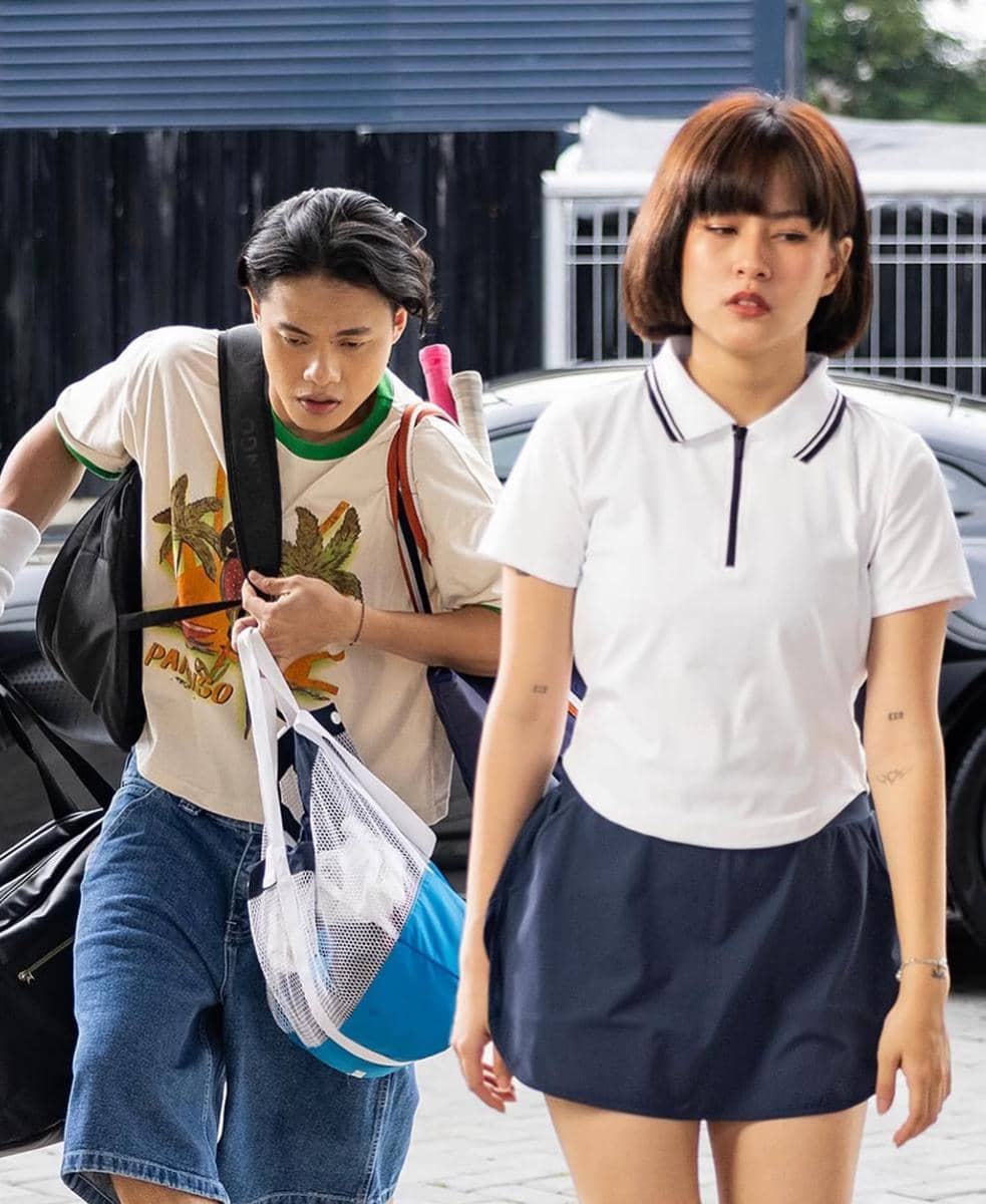 Seorang pria membawa tas olahraga dan raket berjalan di belakang seorang wanita berambut pendek mengenakan pakaian tenis putih dan rok biru.