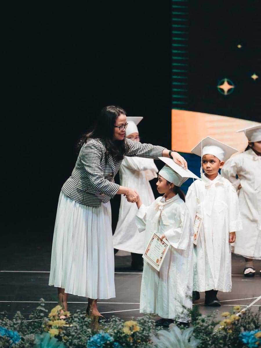 Seorang anak mengenakan toga putih menerima ijazah dari seorang wanita di atas panggung saat acara wisuda taman kanak-kanak.