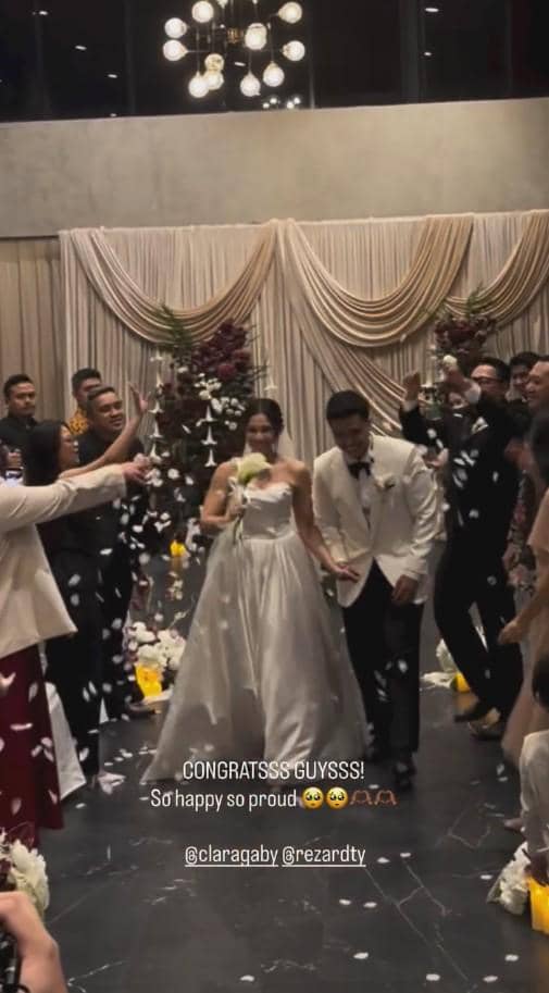Pasangan pengantin berjalan di lorong pernikahan dengan tamu yang menaburkan bunga, suasana bahagia dan meriah di dalam ruangan elegan.