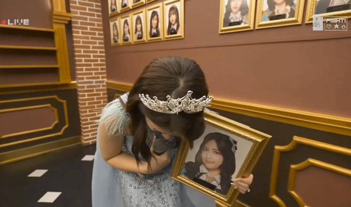 Seorang anggota JKT48 mengenakan gaun biru dan mahkota sambil memegang bingkai foto dirinya di dinding berisi potret anggota lain.