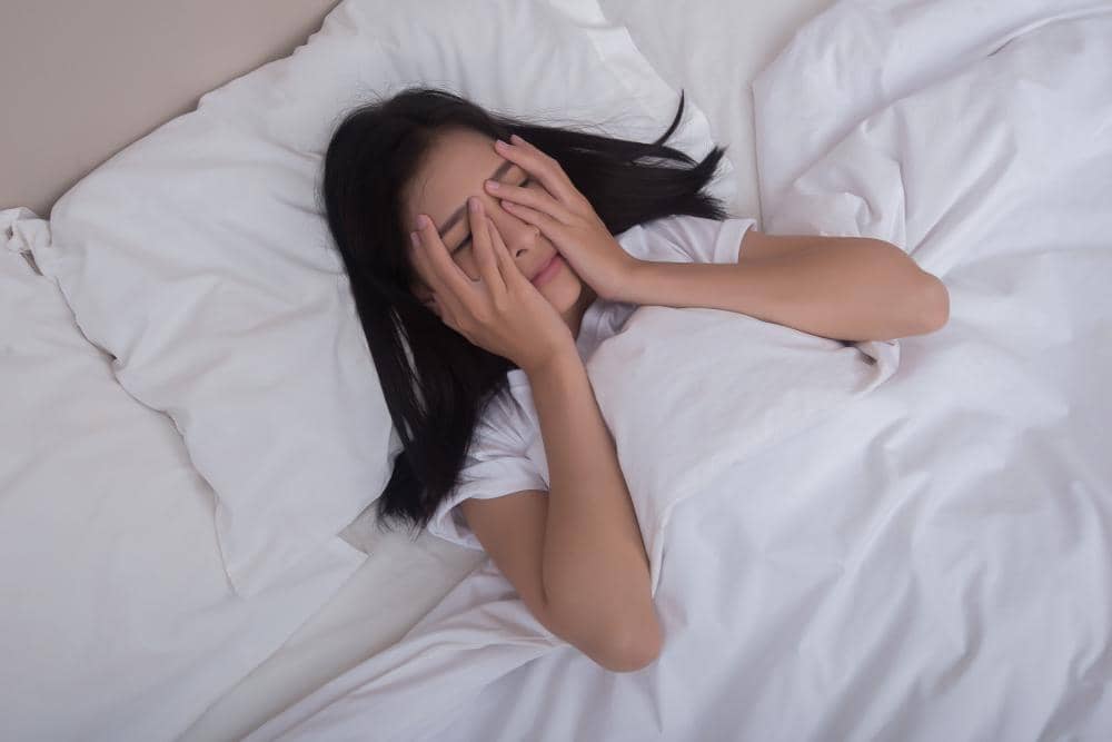Seorang perempuan berbaring di tempat tidur dengan tangan menutupi wajah, tampak lelah dan kurang tidur di kamar bernuansa putih.