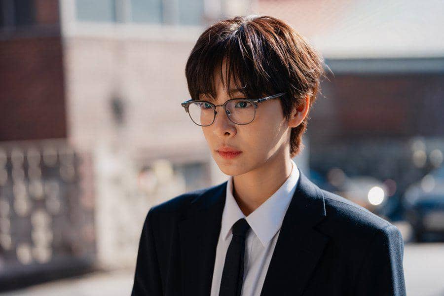 Seorang karakter mengenakan setelan jas hitam dan kacamata berdiri di luar ruangan dengan latar bangunan bata dalam cuplikan drama Love in Disguise.