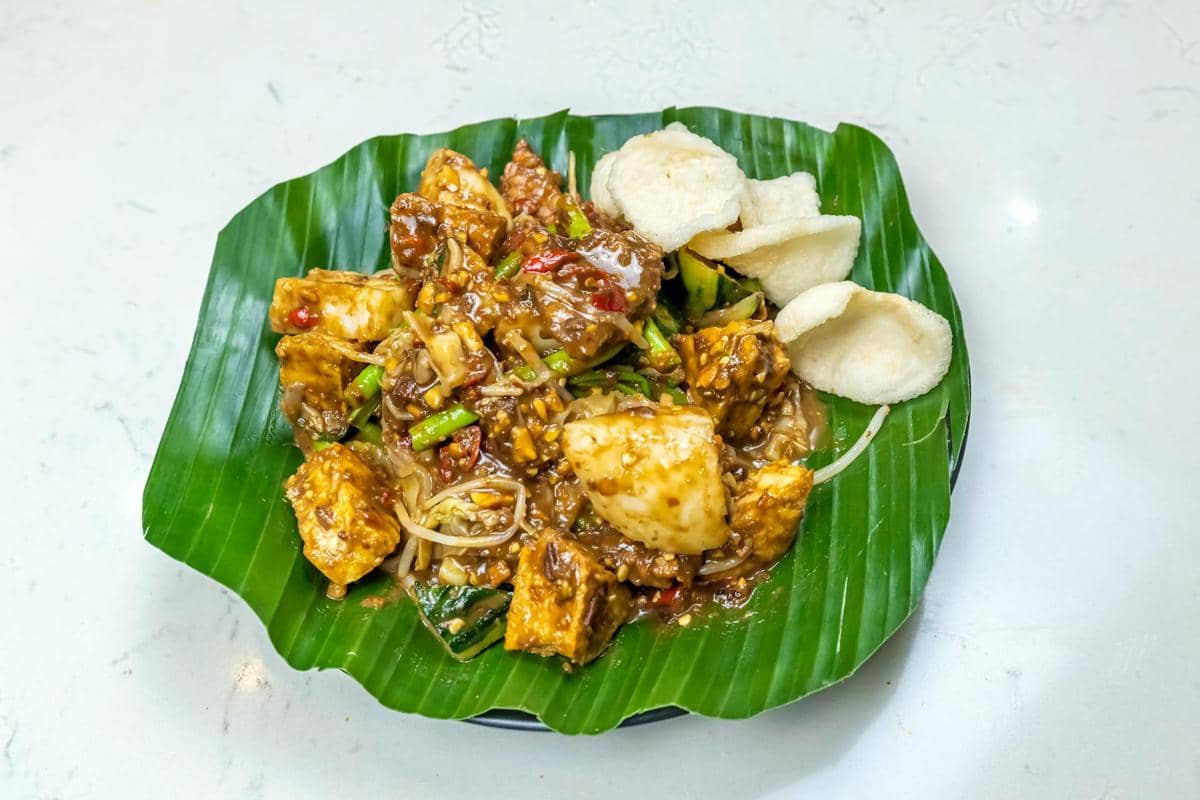 Pecel disajikan di atas daun pisang dengan bumbu kacang, potongan tahu, sayuran rebus, dan kerupuk di sisi piring.
