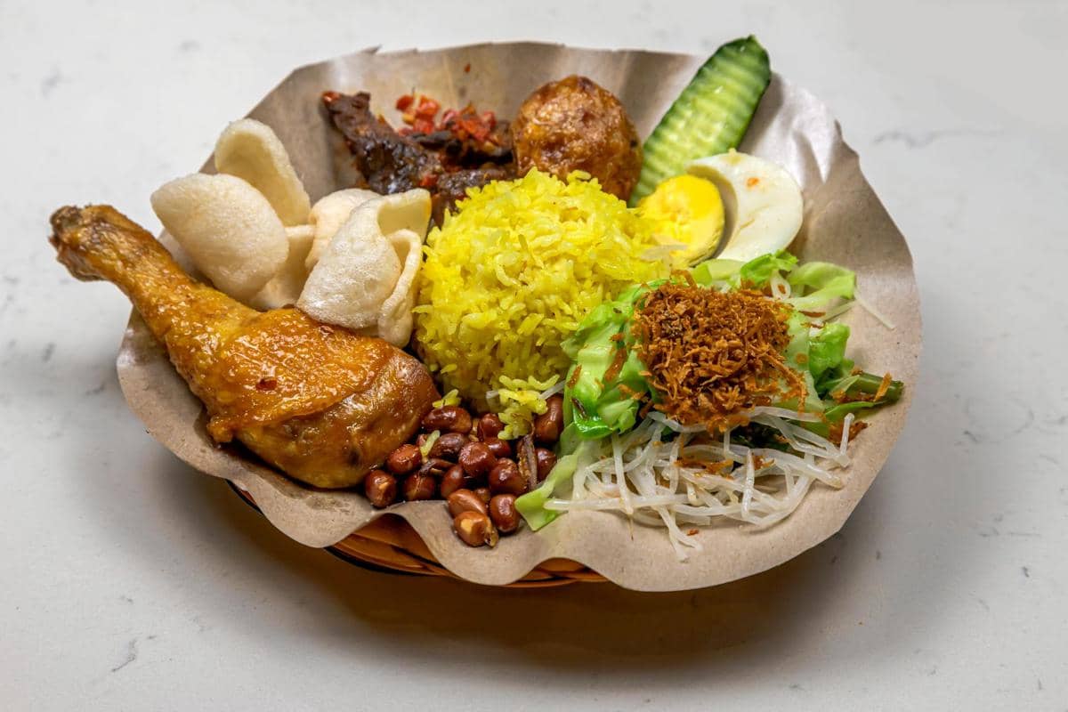 Sepiring nasi kuning lengkap dengan ayam goreng, kerupuk, telur rebus, kacang tanah, sayuran, dan sambal di atas alas kertas.