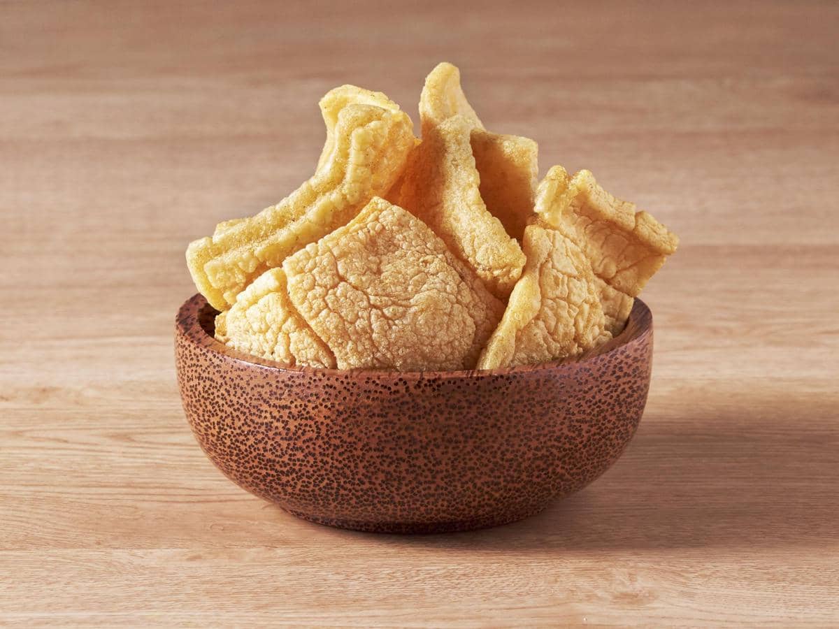 Beberapa potong kerupuk berwarna kuning keemasan dengan tekstur renyah tersaji dalam mangkuk cokelat di atas meja kayu.