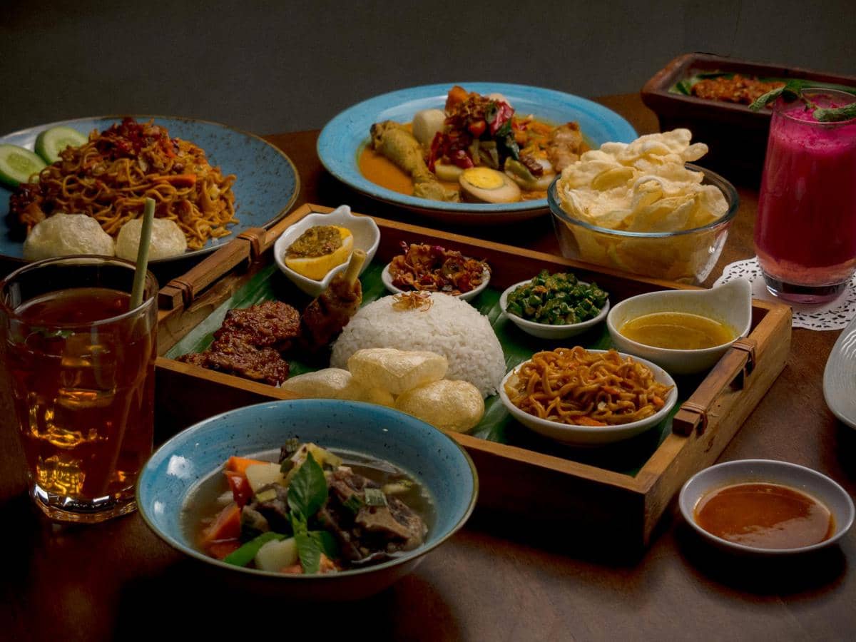 Aneka hidangan khas Indonesia tersaji di meja, termasuk nasi, mi goreng, sate, sayur sop, kerupuk, dan minuman tradisional.