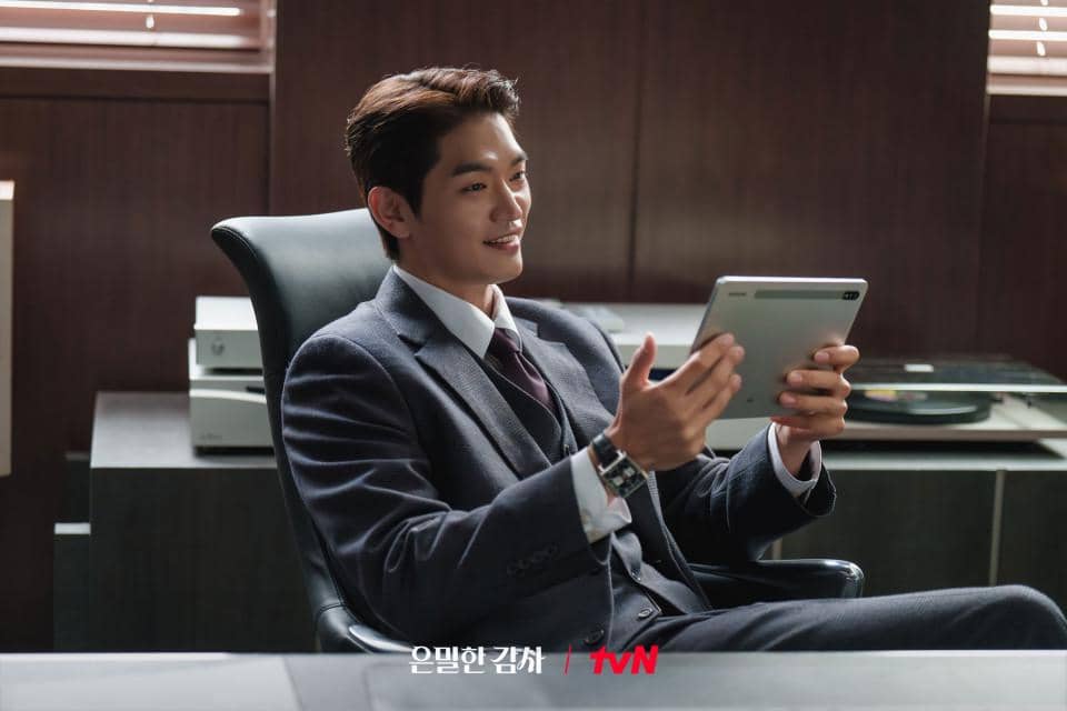 Seorang pria mengenakan setelan jas duduk santai di kursi kantor sambil memegang tablet dan tersenyum di ruangan kerja modern.