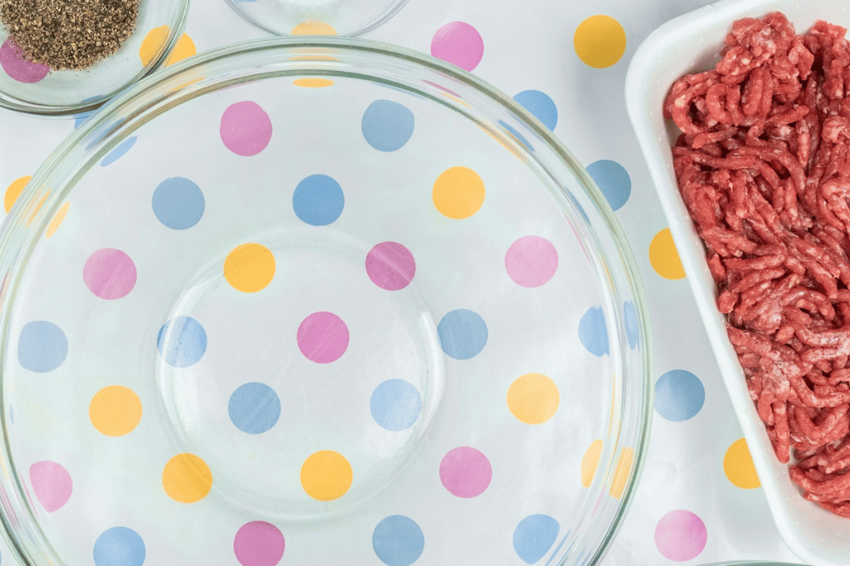 Daging sapi giling mentah dalam wadah putih di samping mangkuk kaca besar di atas meja bermotif polkadot warna-warni.
