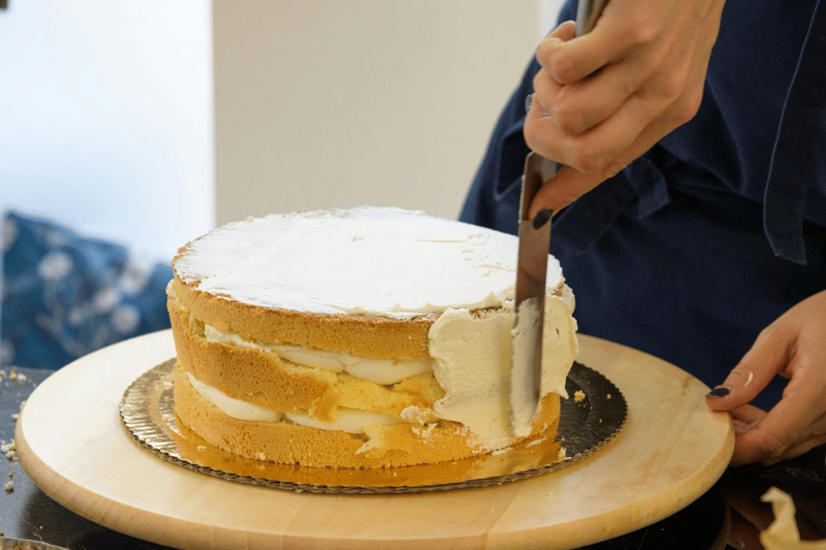 Seseorang sedang mengoleskan krim putih pada kue lapis berwarna kuning menggunakan spatula di atas alas kayu bundar.