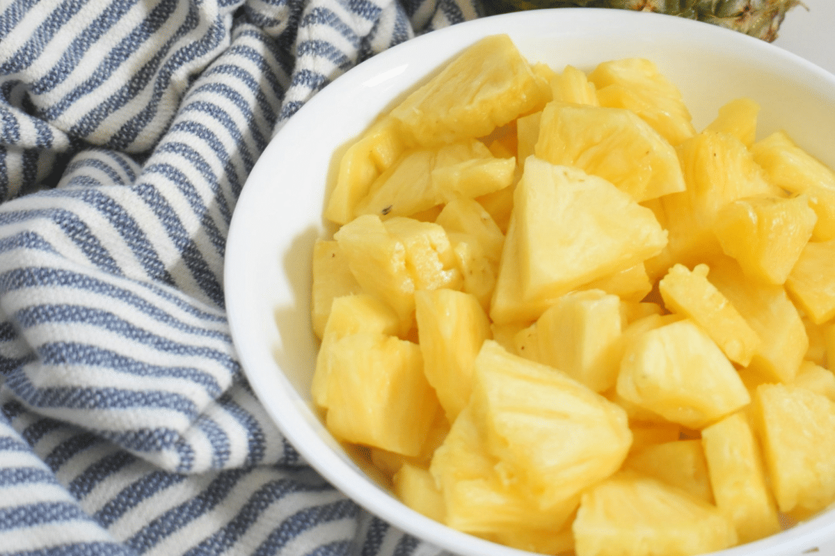 Potongan buah nanas segar berwarna kuning cerah tersaji dalam mangkuk putih di atas kain bergaris biru putih.