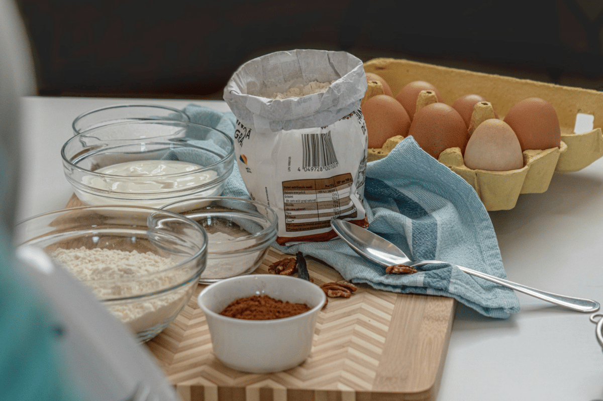 Bahan-bahan membuat roti zukini seperti tepung, telur, yogurt, dan bubuk cokelat tersusun di meja dapur dengan kain biru.
