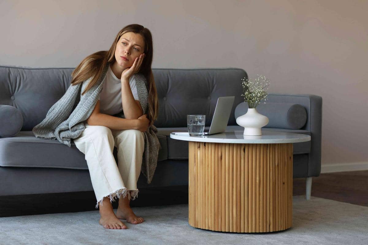 Seorang wanita duduk di sofa abu-abu dengan ekspresi termenung, mengenakan sweater abu dan celana putih di ruang tamu minimalis.