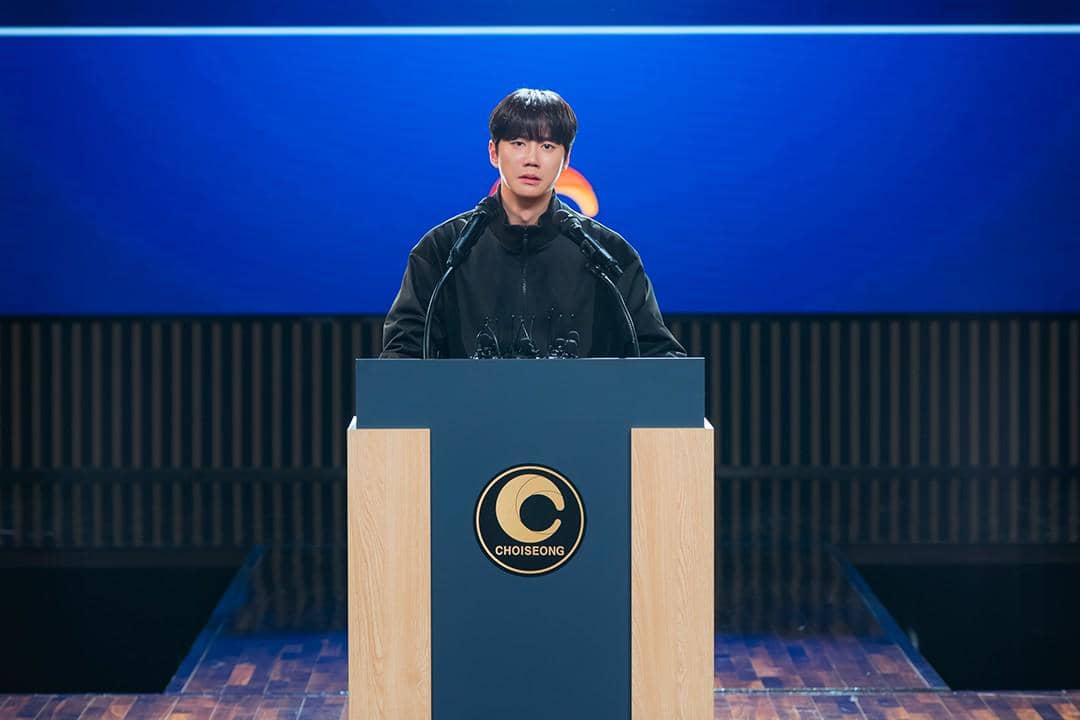Seorang pria berdiri di podium dengan dua mikrofon di depannya dalam adegan drama Reborn Rookie produksi JTBC.