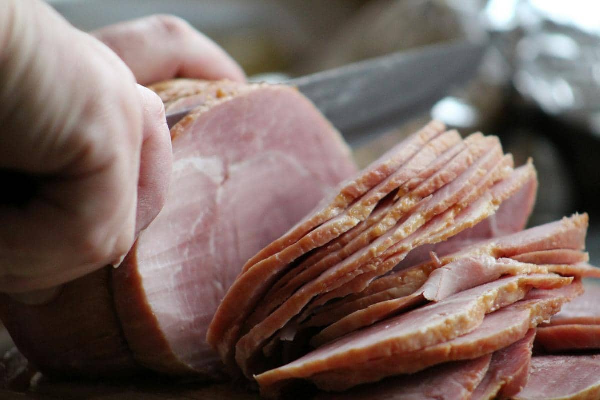 Tangan sedang mengiris ham matang menjadi potongan tipis menggunakan pisau tajam di atas permukaan dapur.