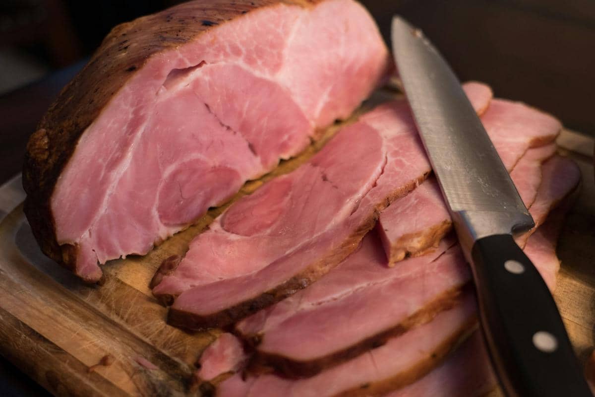 Potongan ham panggang berwarna merah muda di atas talenan kayu dengan pisau dapur di sampingnya siap disajikan.