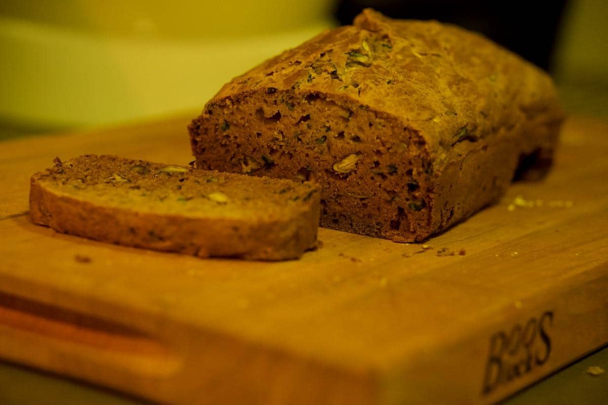 Sebatang roti zukini berwarna cokelat dengan satu potongan di papan kayu, menampilkan tekstur lembut dan potongan sayuran di dalamnya.