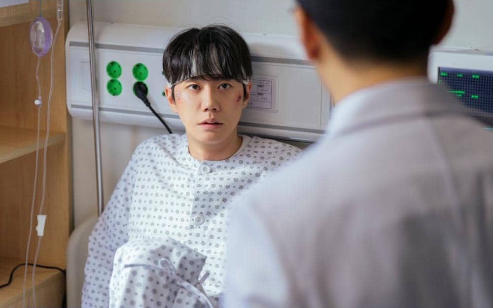 Seorang pria dengan luka di wajah mengenakan pakaian rumah sakit duduk di ranjang sambil berbicara dengan dokter di ruang perawatan.