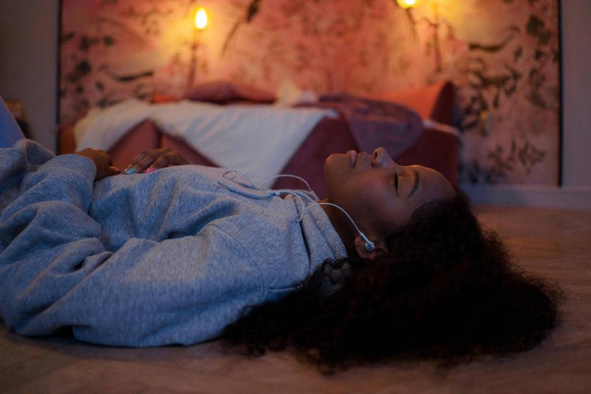 Seorang perempuan berbaring santai di lantai kamar dengan earphone di telinga, menikmati musik dalam suasana hangat dan tenang.