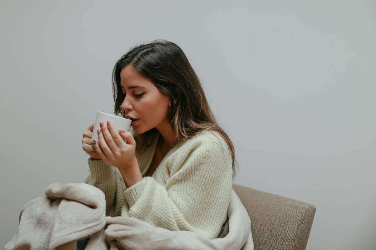 Seorang wanita mengenakan sweater krem duduk di kursi sambil memegang cangkir putih dan menikmati minuman hangat dengan selimut di pangkuannya.
