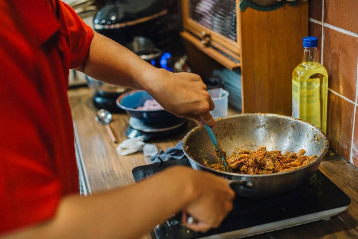 Seseorang mengenakan baju merah sedang mengaduk masakan berbumbu di wajan di atas kompor di dapur rumah.