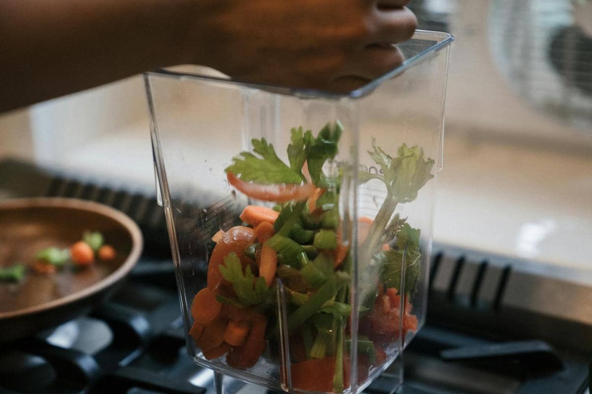 Tangan memasukkan potongan sayuran segar seperti wortel dan seledri ke dalam blender di dapur untuk dihaluskan.