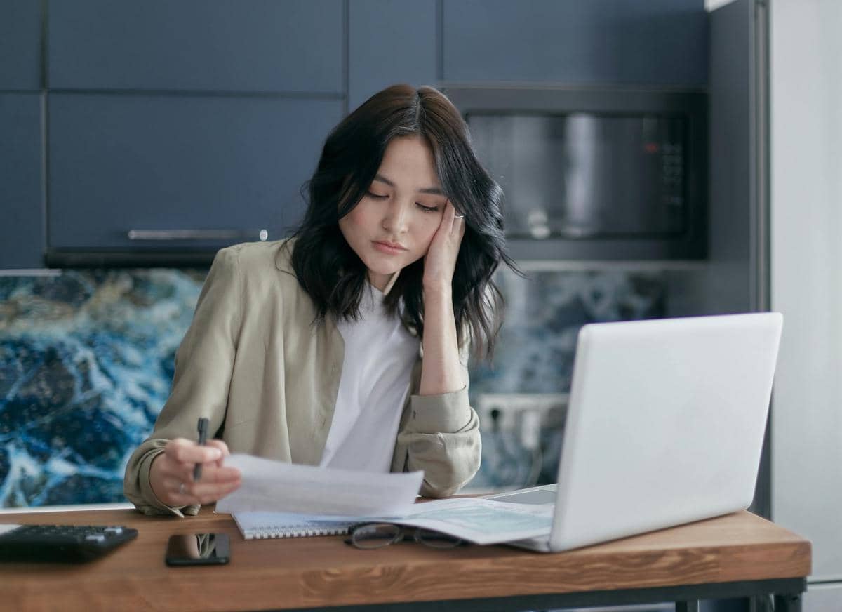 Seorang wanita tampak lelah sambil membaca dokumen di meja kerja dengan laptop terbuka di depannya di ruang dapur modern.