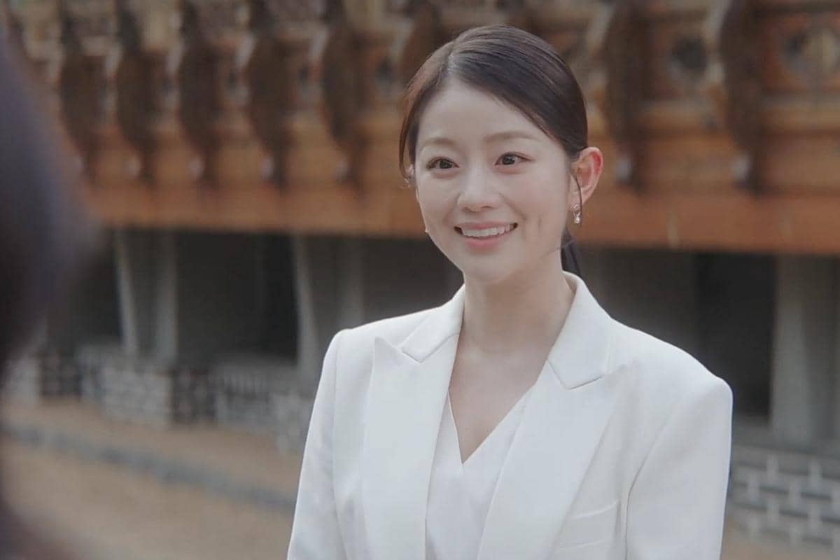 Seorang wanita mengenakan setelan jas putih tersenyum hangat di luar ruangan dengan latar bangunan bergaya tradisional Korea.