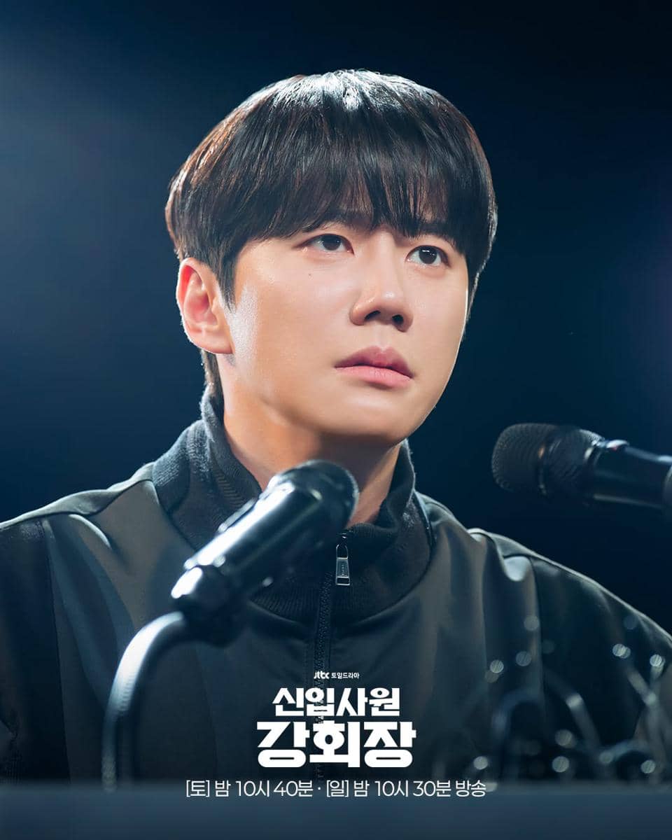 Seorang pria mengenakan jaket hitam duduk di depan mikrofon dengan ekspresi serius dalam adegan drama Reborn Rookie.