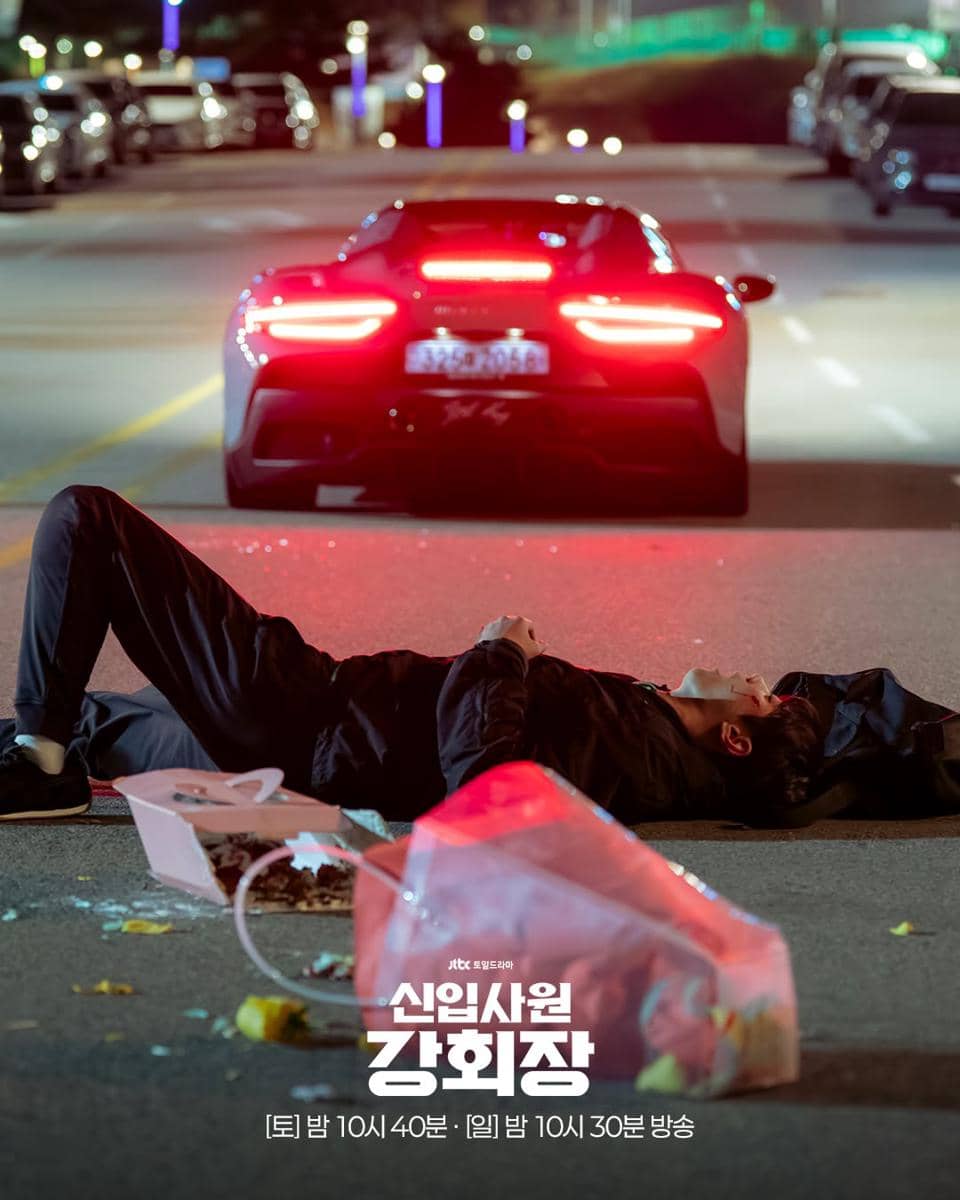 Seorang pria terbaring di jalan pada malam hari di depan mobil sport merah dengan lampu belakang menyala terang.