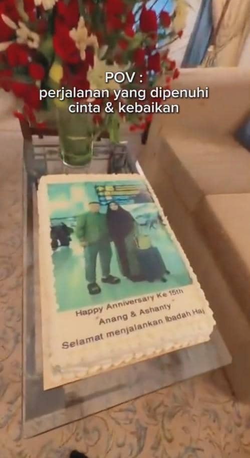 Kue ulang tahun pernikahan ke-15 dengan foto pasangan di atas meja kaca, dihiasi bunga merah di vas besar di latar belakang.